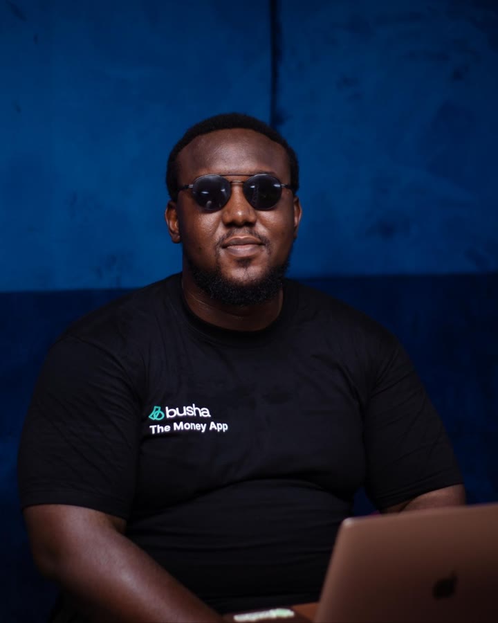 Raji Rufai in a black Busha T-shirt and round sunglasses, seated at a laptop